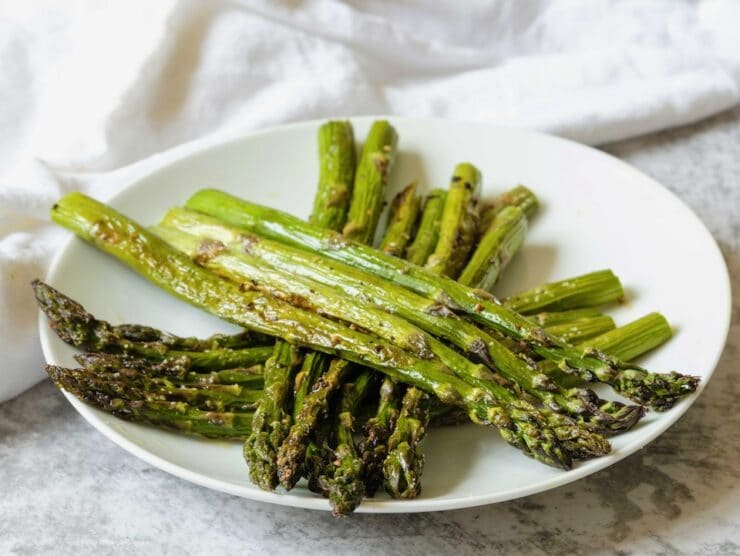Three small piles of aligned roasted asparagus criss-crossed diagonally on a white plate, linen napkin in background.