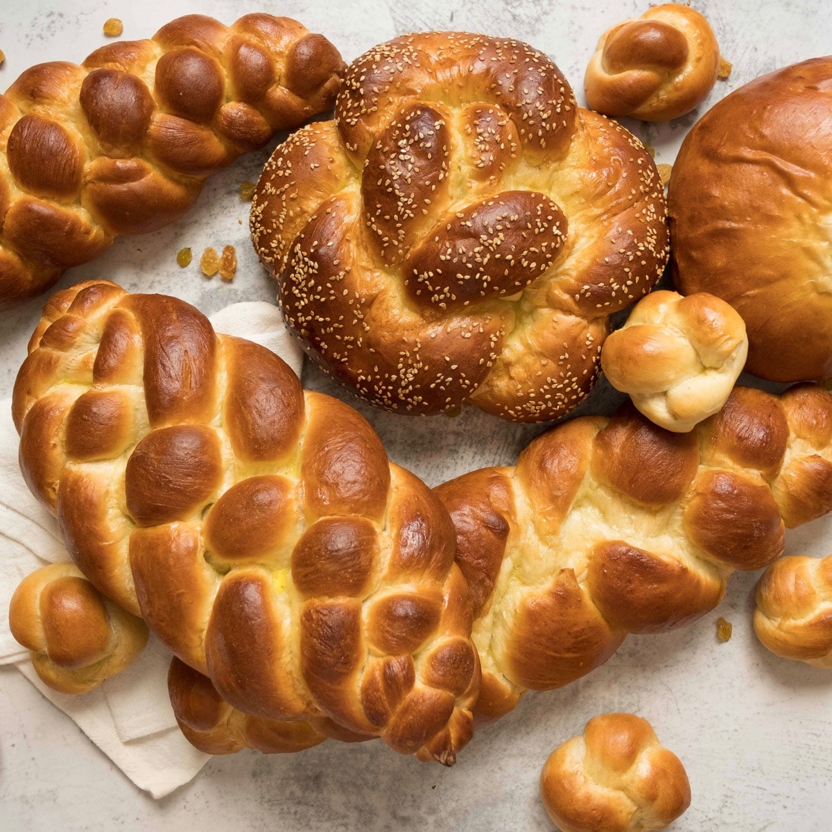 Square Crop - Overhead shot - multiple challah braids - three strand, four strand, round braid, turban challah, and challah rolls, on marble countertop with cloth napkin beneath.
