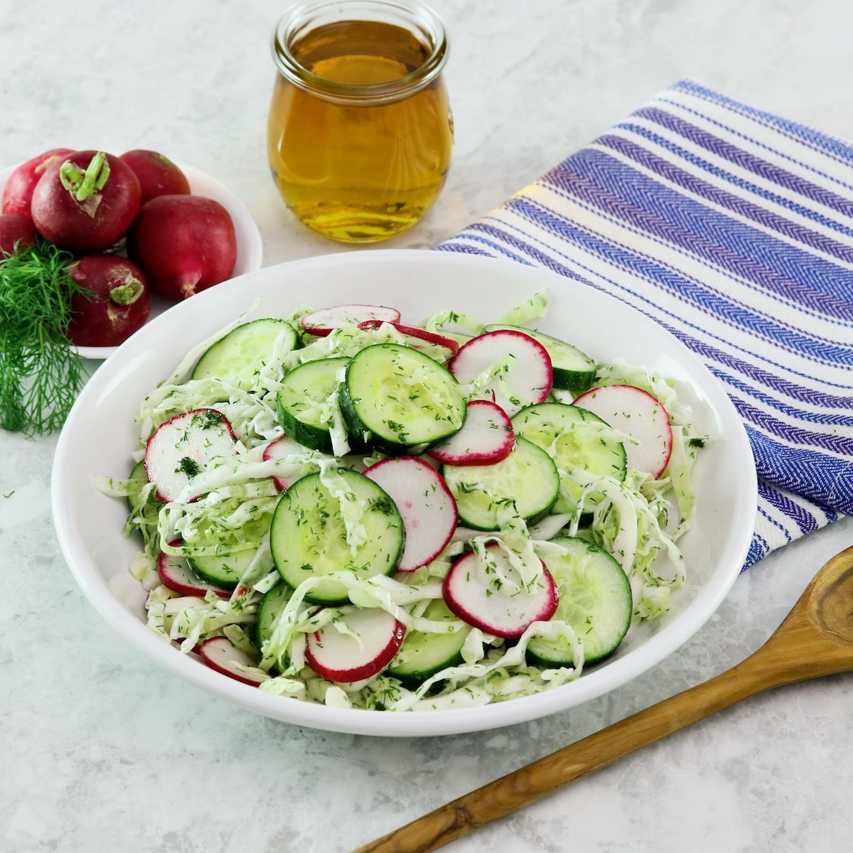 Close up shot of a large bowl filled with crunch pickled salad. A wooden bowl and blue striped napkin sit off to the right side. To the left there is a bunch of fresh radishes.