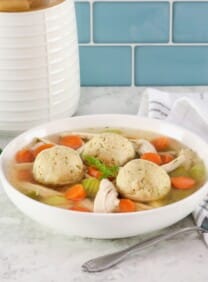Matzo ball soup - horizontal shot of classic Jewish chicken soup with fluffy matzo balls on a white countertop. Spoon and linen napkin beside the bowl, fresh green herbs and white ceramic jar in background.