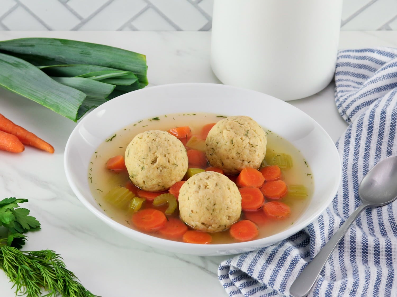Horizontal shot of floater matzo balls in a shallow bowl of vegetarian matzo ball soup with carrot slices, pieces of celery, and golden saffron broth. Spoon, fresh vegetables, and linen napkin on the white marble counter beside the bowl. Tiles and white jar in background.