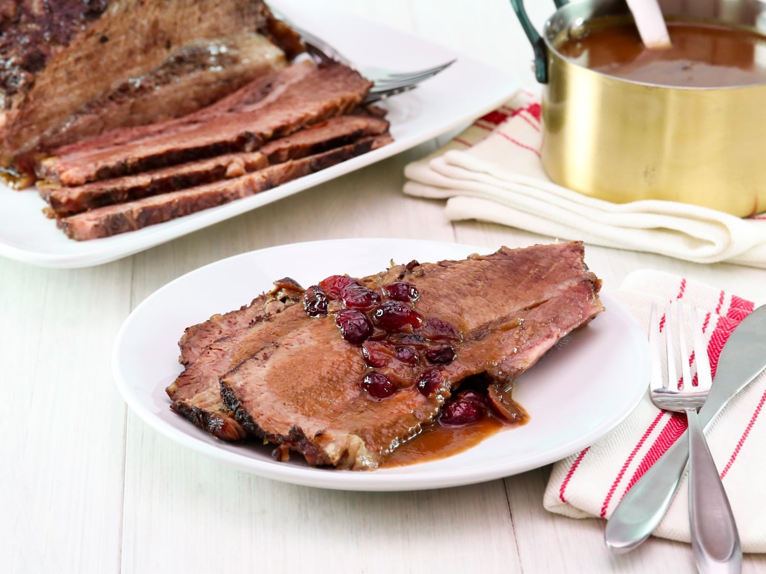 Horizontal shot - white wooden table set with dinner plate of Cranberry Chipotle brisket slices slathered in cranberry sauce and juices, with cloth napkin and utensils. Platter of sliced brisket and golden pan of sauce on a cloth napkin in the background.