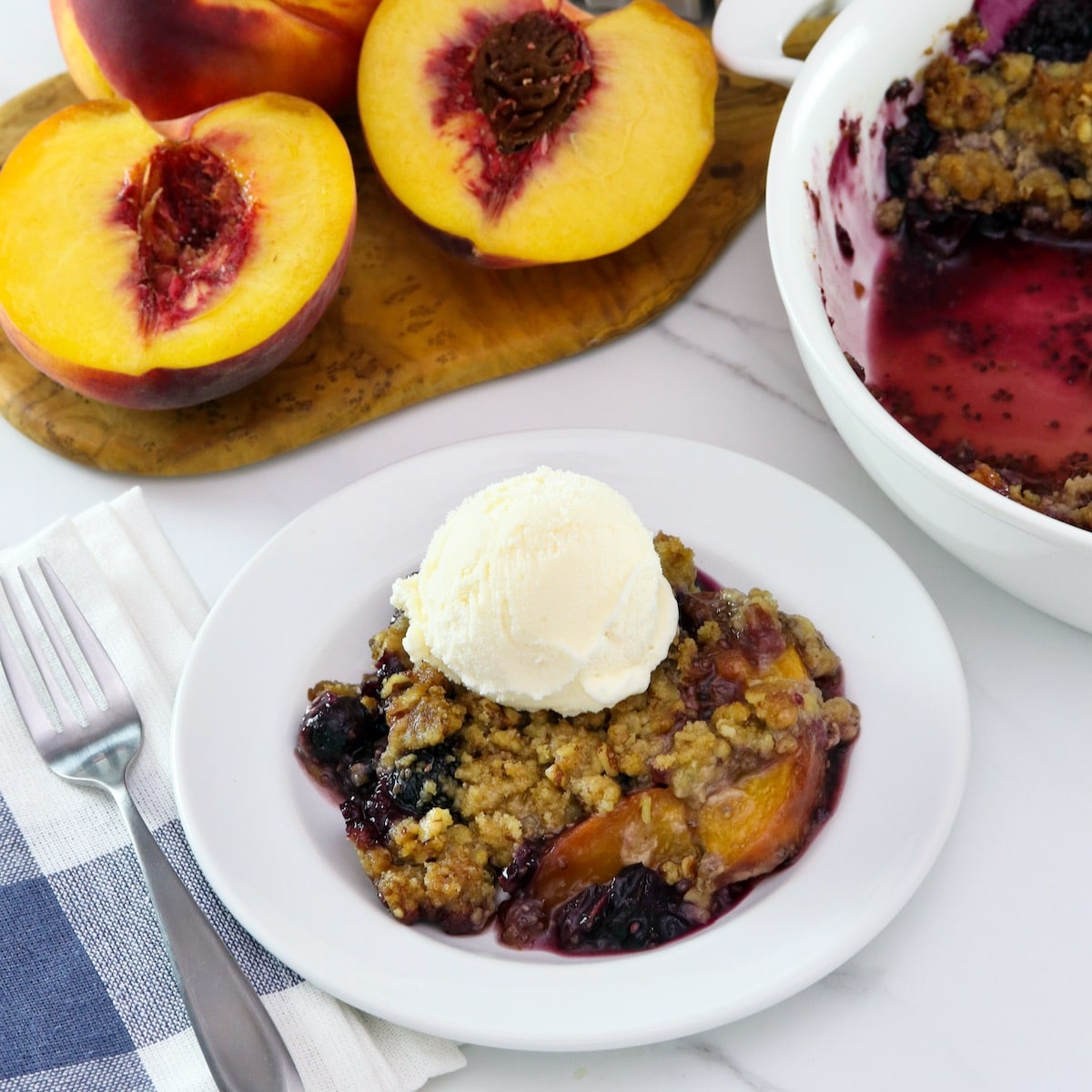 Square image - peach and blueberry crisp on a small white plate, topped with a scoop of vanilla ice cream. A white baking dish of the remaining crisp sits off to the right next to a pile of fresh peaches.