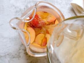 Overhead shot of clear simple syrup being poured into a glass pitcher with sliced peaches at the bottom.