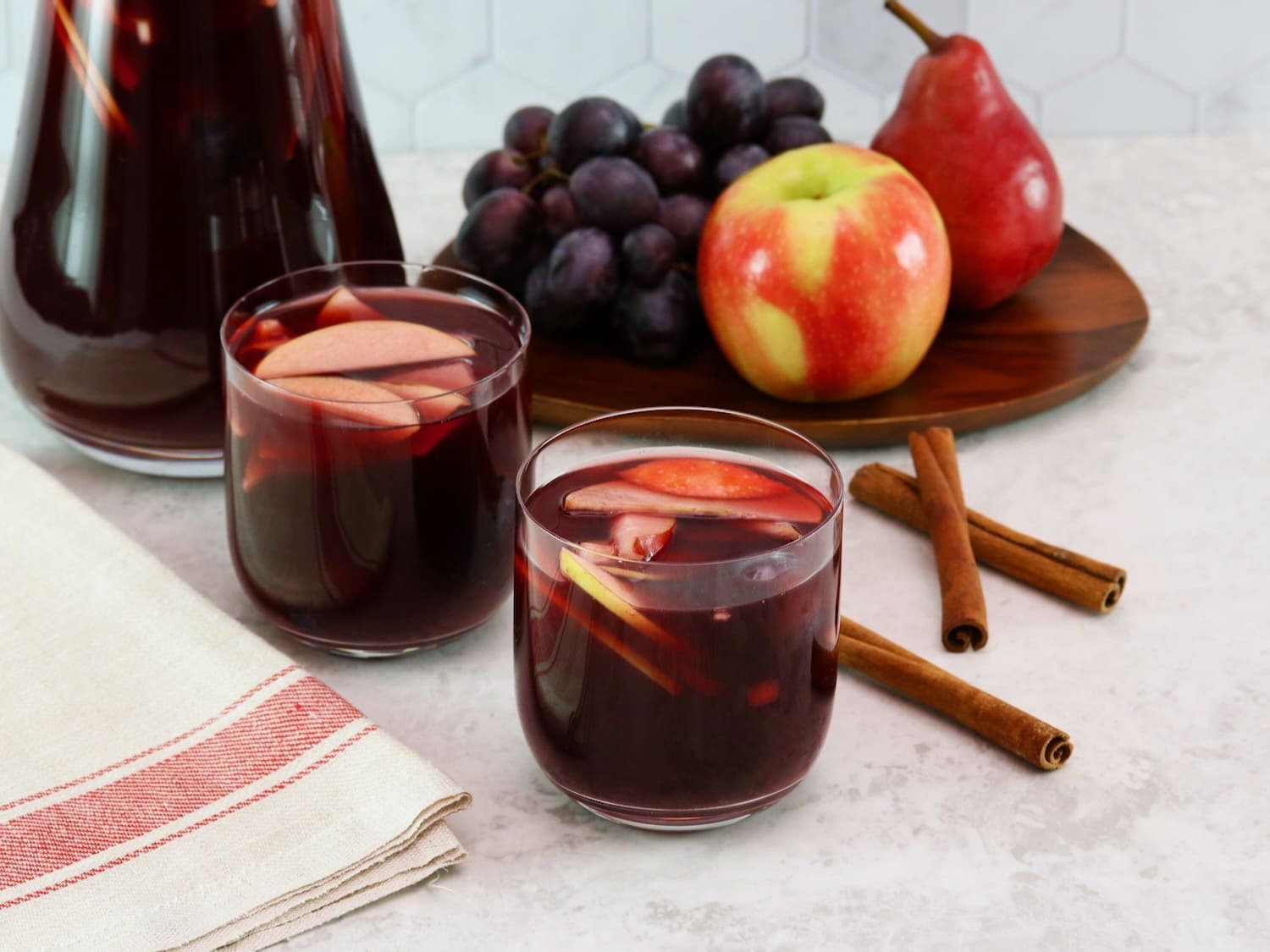 Close up shot - two unstemmed wine glasses filled with red wine Thanksgiving sangria, fresh fruit slices - apples and grapes floating in the cups. Linen towel beside the cups. On a white marble countertop with three cinnamon sticks, a platter of fruit - apple, grapes, pears - and a large pitcher of sangria in the background.