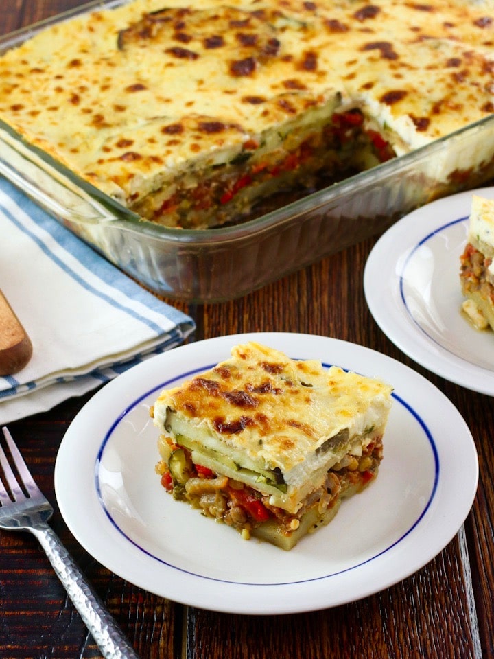 Slice of Roasted Vegetable Moussaka on a small plate in front of casserole dish with moussaka, fork, cloth napkin and serving utensil on wooden tabletop.