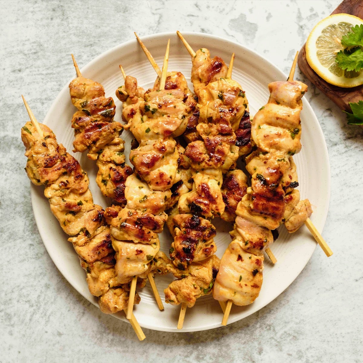 Overhead square shot - plate of Lemony Marinated Chicken Skewers on countertop, lemon and parsley garnish on wooden cutting board beside the plate.
