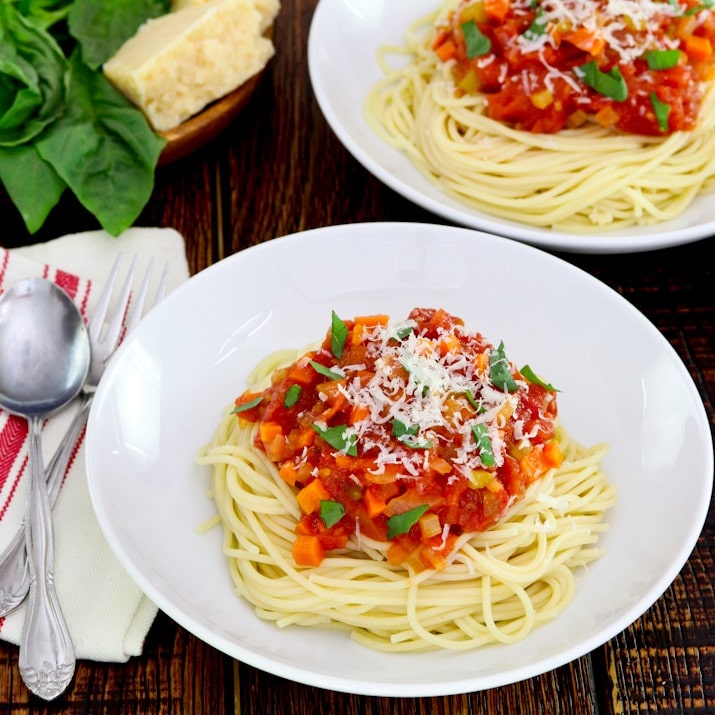 Square crop - plate of spaghetti pasta with pomodoro sauce topped with basil and parmesan, cloth napkin with utensils beside it, another plate of pasta, parmesan block and fresh basil in background.