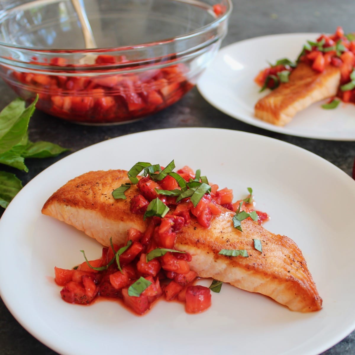 Square Crop - seared salmon fillet topped with macerated strawberry balsamic sauce and fresh basil chiffonade, another plate of the same entree in background, with a glass bowl of macerating strawberries and a pile of fresh basil in the back.
