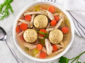 Overhead shot of a bowl of floater matzo balls in a shallow bowl of Jewish chicken soup with carrot slices, pieces of celery, and golden broth. Spoon, fresh herbs, and linen napkin on the white marble counter beside the bowl.